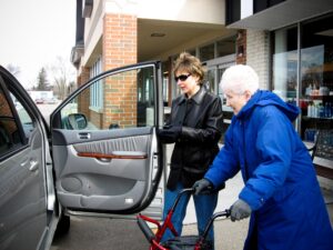 A woman is opening the car door for an older woman with a walker to enter.
