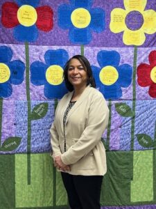 Woman with dark hair in a off white sweater posing in front of a colorful floral quilt.